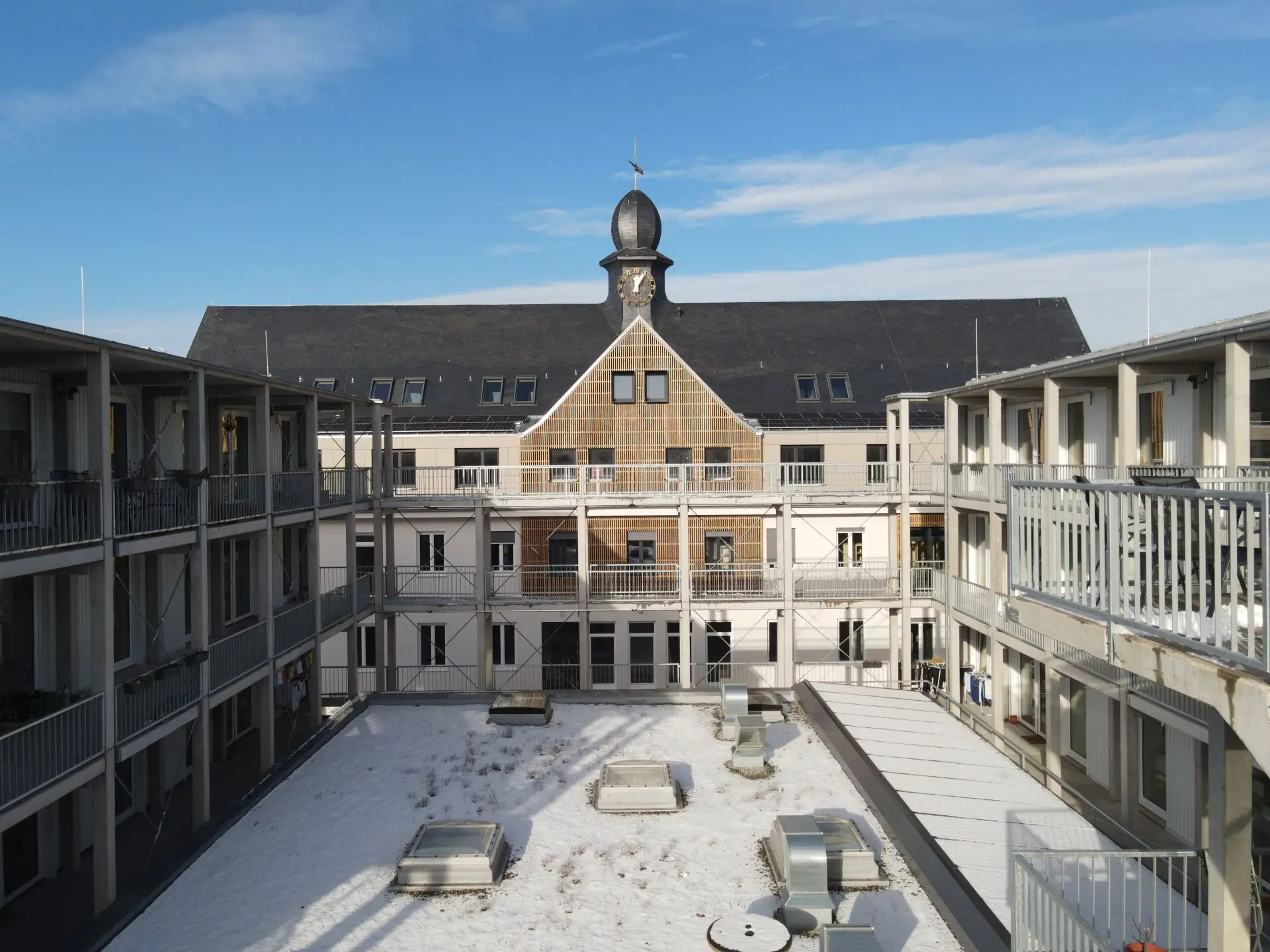 Das Foto zeigt einen Gebäudekomplex mit Laubengängen. Man blickt in Zentalperspektive auf ein Gebäude mit schwarzem Satteldach, auf dem mittig ein kleines Türmchen mit Uhr sitzt, direkt über einem mit Holz verkleideten Zwerchgiebel. Die Gebäudeteile rechts und links davon wirken moderner mit Flachdächern und Betonstützen.