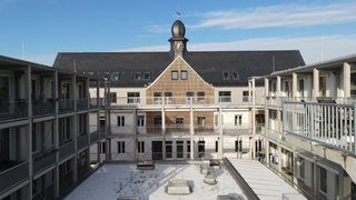 Das Foto zeigt einen Gebäudekomplex mit Laubengängen. Man blickt in Zentalperspektive auf ein Gebäude mit schwarzem Satteldach, auf dem mittig ein kleines Türmchen mit Uhr sitzt, direkt über einem mit Holz verkleideten Zwerchgiebel. Die Gebäudeteile rechts und links davon wirken moderner mit Flachdächern und Betonstützen. Kachel Vorschau