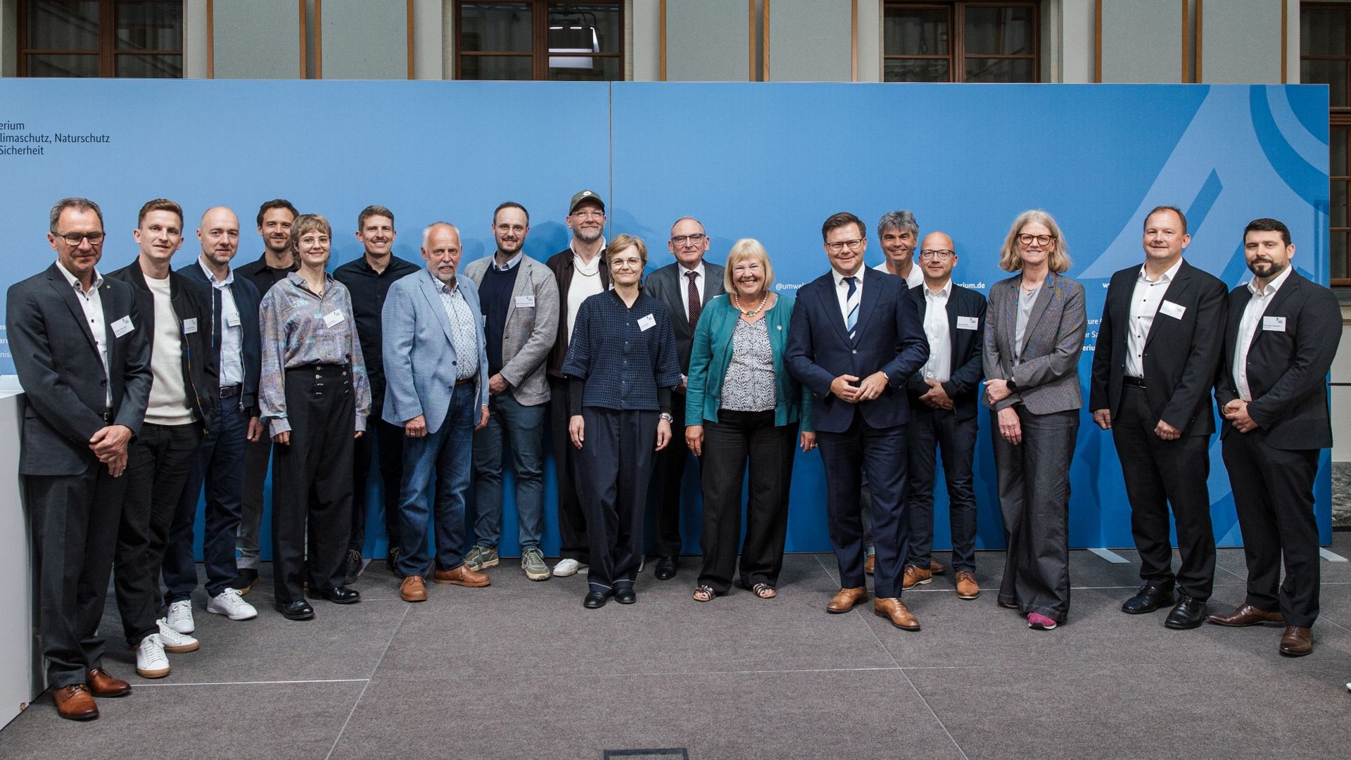 Gruppenfoto mit 18 gut gekleideten Personen vor einer blauen Rückwand.