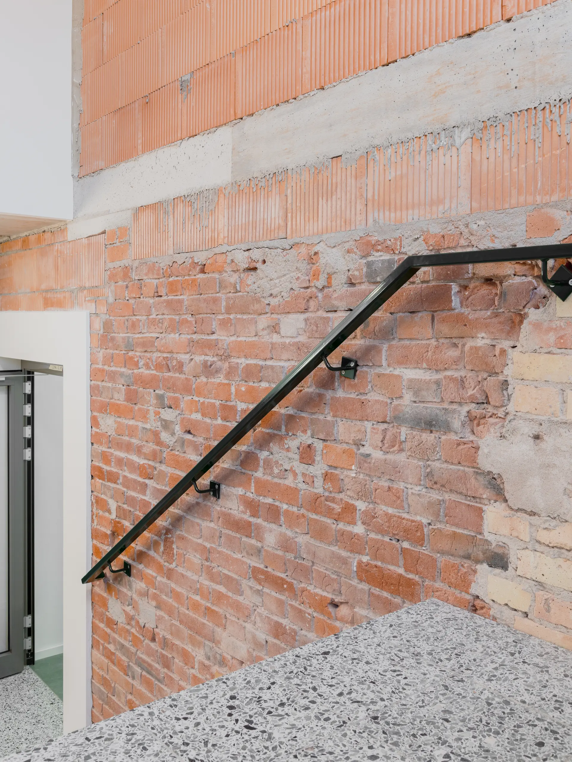 Man sieht eine Wand in einem Treppenhaus, sie ist unverputzt und besteht aus einem Mix von alten und neuen Ziegeln in verschiedenen Formaten und Farben.