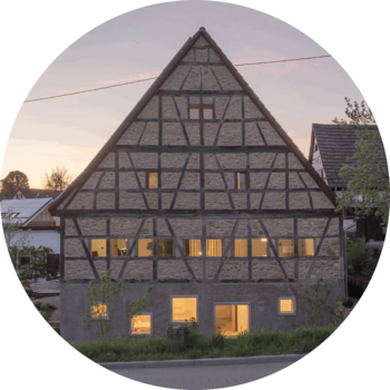 Das Foto zeigt die Giebelseite einer alten Fachwerkscheune. Das Erdgeschoss hebt sich in der Gestaltung von dem Rest des Gebäudes ab, es ist aus Sichtbeton mit 5 quadratischen Fenstern in unterschiedlichen Größen, die versetzt angeordnet sind.