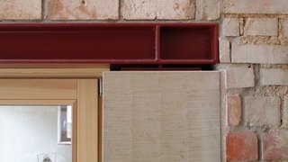 Auf dem Foto sieht man ein Konstruktionsdetail. Ein Fenster mit Holzrahmen trifft auf einen roten Stahlträger, der den Fenstersturz bildet. Neben dem Fenster ist ein Sandsteinelement, dann beginnt die Backsteinwand. Kachel Vorschau