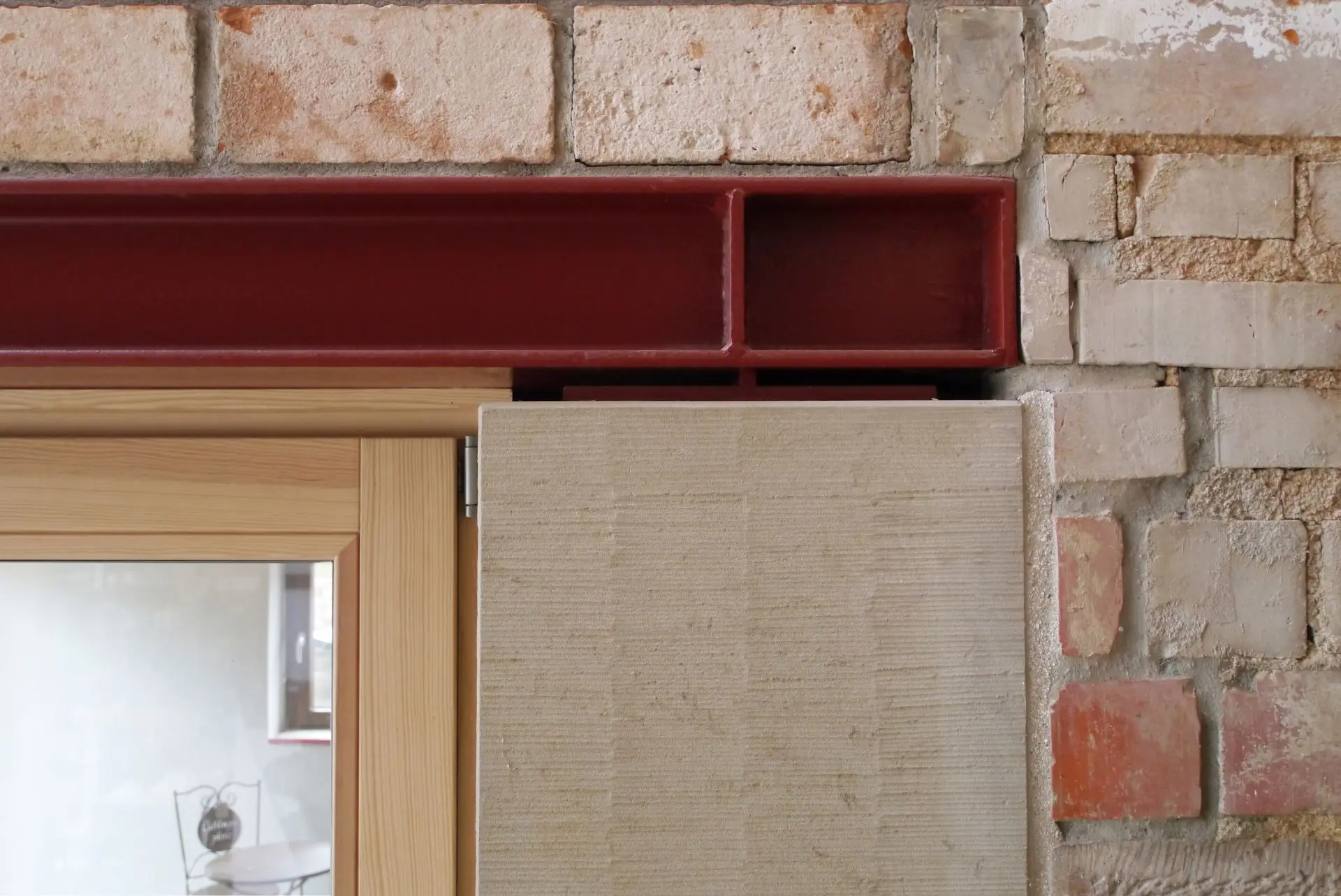 Auf dem Foto sieht man ein Konstruktionsdetail. Ein Fenster mit Holzrahmen trifft auf einen roten Stahlträger, der den Fenstersturz bildet. Neben dem Fenster ist ein Sandsteinelement, dann beginnt die Backsteinwand.