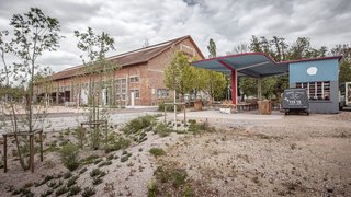 Das Foto zeigt die lange rote Backsteinhalle mit dem Schriftzug “REITHALLE” am Giebel von außen. Auf einem Platz rechts neben der Halle steht ein blauer Kiosk mit einem überdachten Vorplatz, auf dem viele Tische und Sitzbänke stehen. Junge Bäume wurden auf dem umliegenden Areal gepflanzt. Kachel Vorschau