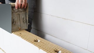 Auf dem Foto ist zu sehen, wie ein brauner Mörtel mit einer Mörtelrolle auf eine Mauer aus Porenbetonsteinen aufgetragen wird. Kachel Vorschau