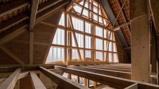 Das Foto zeigt den Dachraum einer Scheune mit Blick Richtung Giebel. Die Dachkonstruktion besteht aus älteren dunklen und neuen hellen Balken. Das Fachwerk des Giebels ist nur an den Seiten mit Backstein ausgefacht, im mittlernen Bereich wurde das Fachwerk offengelegt und von außen mit einer halbtransparenten Verkleidung versehen, die Licht in den Innenraum fallen lässt. Kachel Vorschau