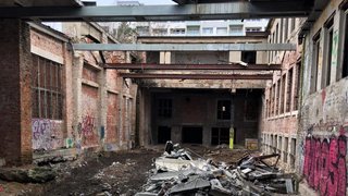 Das Foto zeigt eine verfallene Industriehalle aus Backstein mit vergitterten Fenstern, auf dem Boden liegt ein Haufen Bauabfälle. Die Wände sind voller Graffiti. Das Dach wurde entfernt, nur noch Stahlträger überspannen den Raum, der jetzt eher ein Hof ist. Durch die Öffnung sieht man den Himmel und im Hintergrund überragt ein Plattenbau das Gebäude.