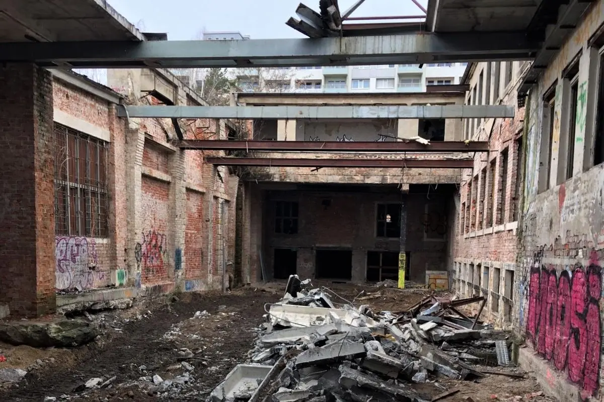 Das Foto zeigt eine verfallene Industriehalle aus Backstein mit vergitterten Fenstern, auf dem Boden liegt ein Haufen Bauabfälle. Die Wände sind voller Graffiti. Das Dach wurde entfernt, nur noch Stahlträger überspannen den Raum, der jetzt eher ein Hof ist. Durch die Öffnung sieht man den Himmel und im Hintergrund überragt ein Plattenbau das Gebäude.