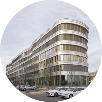 Das Foto zeigt ein Bürogebäude mit abgerundeten Ecken und einer geschwungenen Fassade. Das Gebäude hat 6 Geschosse und ist durch eine Bandfassade in horizontale Fensterbänder und hellgraue Verkleidung gegliedert. Im Vordergrund sind geparkte Autos und ein Fußgänger zu sehen, es stehen junge Bäume entlang des Gebäudes.