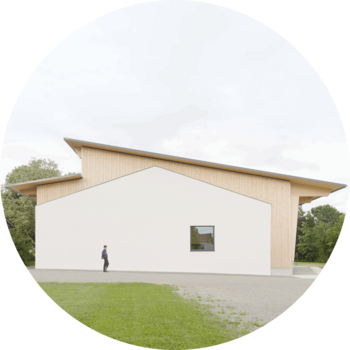 Das Foto zeigt die Stirnseite einer Mehrzweckhalle mit einer zusammengesetzten Fassade. Der eine Teil ist weiß verputzt und hat die Form eines eingeschossigen Baus mit Satteldach. Diese Form wird umgeben von einem zweiten mit Holz verkleideten Teil, der in einem versetzten Pultdach endet.