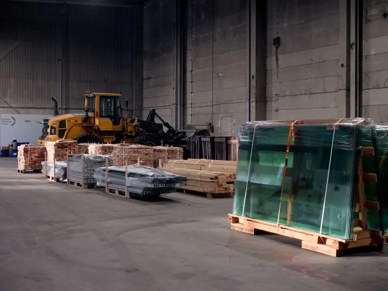 In einer großen Halle lagern verschiedene Baumaterialien auf Paletten, zum Beispiel Ziegel, Holz und Glas.