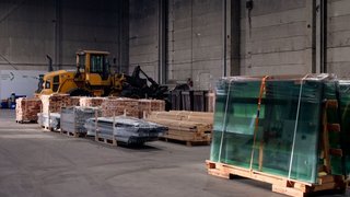 In einer großen Halle lagern verschiedene Baumaterialien auf Paletten, zum Beispiel Ziegel, Holz und Glas. Kachel Vorschau