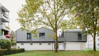Die Aufstockung der Garagenanlage liegt zwischen Mehrfamilienhäusern in einer grünen Umgebung mit Bäumen. Kachel Vorschau