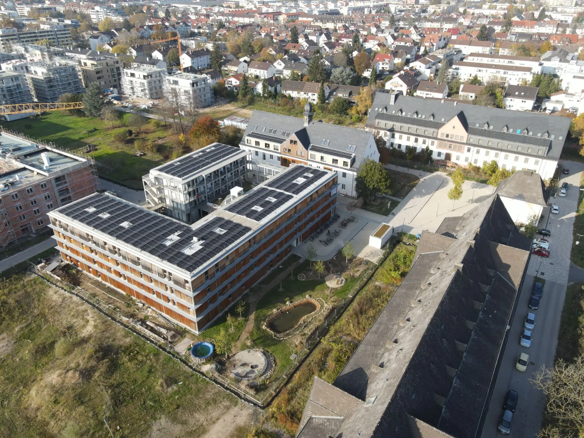 Das Foto ist eine Drohnenaufnahme eines Gebäudekomplexes. Auf den Flachdächern sind Photovoltaikanlagen installiert. Neben den Gebäuden liegt ein Garten mit einem kleinen Teich, einem Pool und jungen Bäumen.