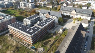 Das Foto ist eine Drohnenaufnahme eines Gebäudekomplexes. Auf den Flachdächern sind Photovoltaikanlagen installiert. Neben den Gebäuden liegt ein Garten mit einem kleinen Teich, einem Pool und jungen Bäumen. Kachel Vorschau