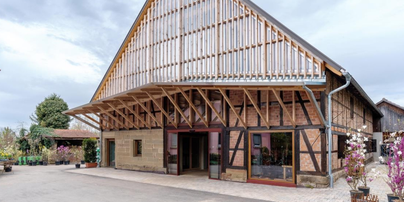 Eine Fachwerkscheune mit Satteldach. Mittig in der Giebelfassade findet sich ein großzügiger Eingangsbereich. Darüber über die ganze Breite des Gebäudes ein freitragendes Vordach. Oberhalb ist die Giebelfassade bis zum First verglast.