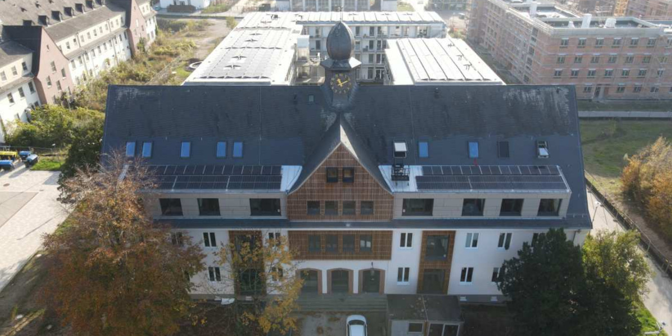 Luftbild eines dreigeschossigen Gebäudes mit Satteldach. Die Fassade ist hell verputzt. Mittig über dem Eingangsbereich wird das dunkle Satteldach von einer Gaube mit orthogonal ausgerichtetem Satteldach durchbrochen. Über der Gaube sitzt ein Türmchen auf dem First, das eine Turmuhr trägt.