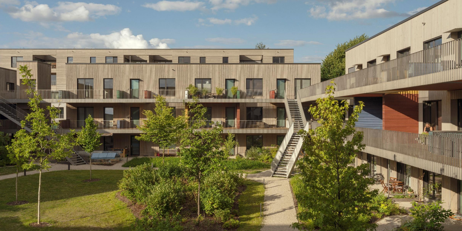 Begrünter Innenhof. Gegenüber und rechts stehen zwei kubische, dreigeschossige Baukörper mit einer Holzfassade. Vor den Fenstern sind Laubengänge angeordnet. In der Bildmitte führt eine Außentreppe zu den Ebenen.
