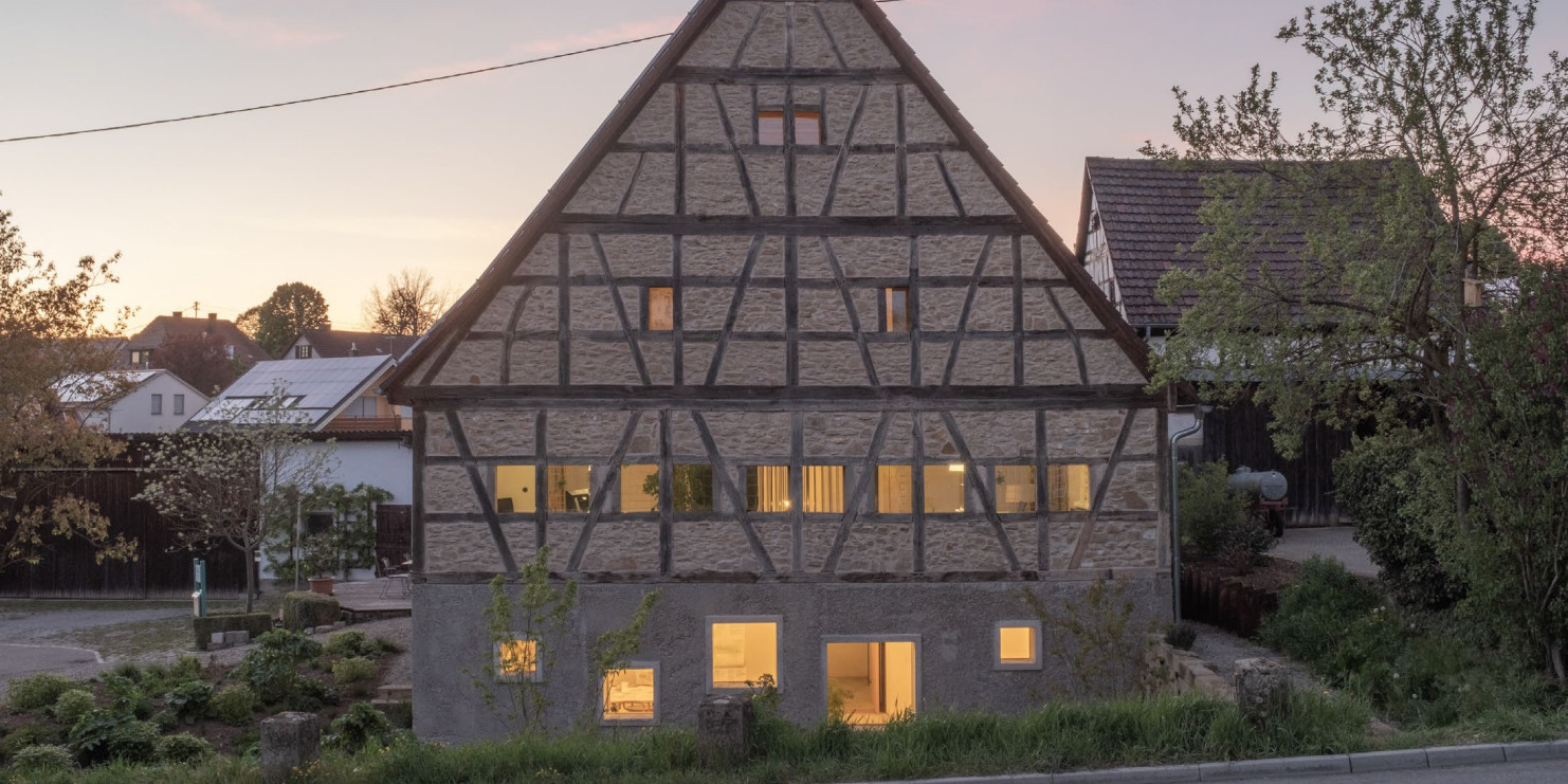 Giebelwand eines viergeschossigen Fachwerkhauses mit Gefachen aus Feldstein und Satteldach. In der dörflichen Szene während der Dämmerung sind die Fenster mit warmem Licht erleuchtet.