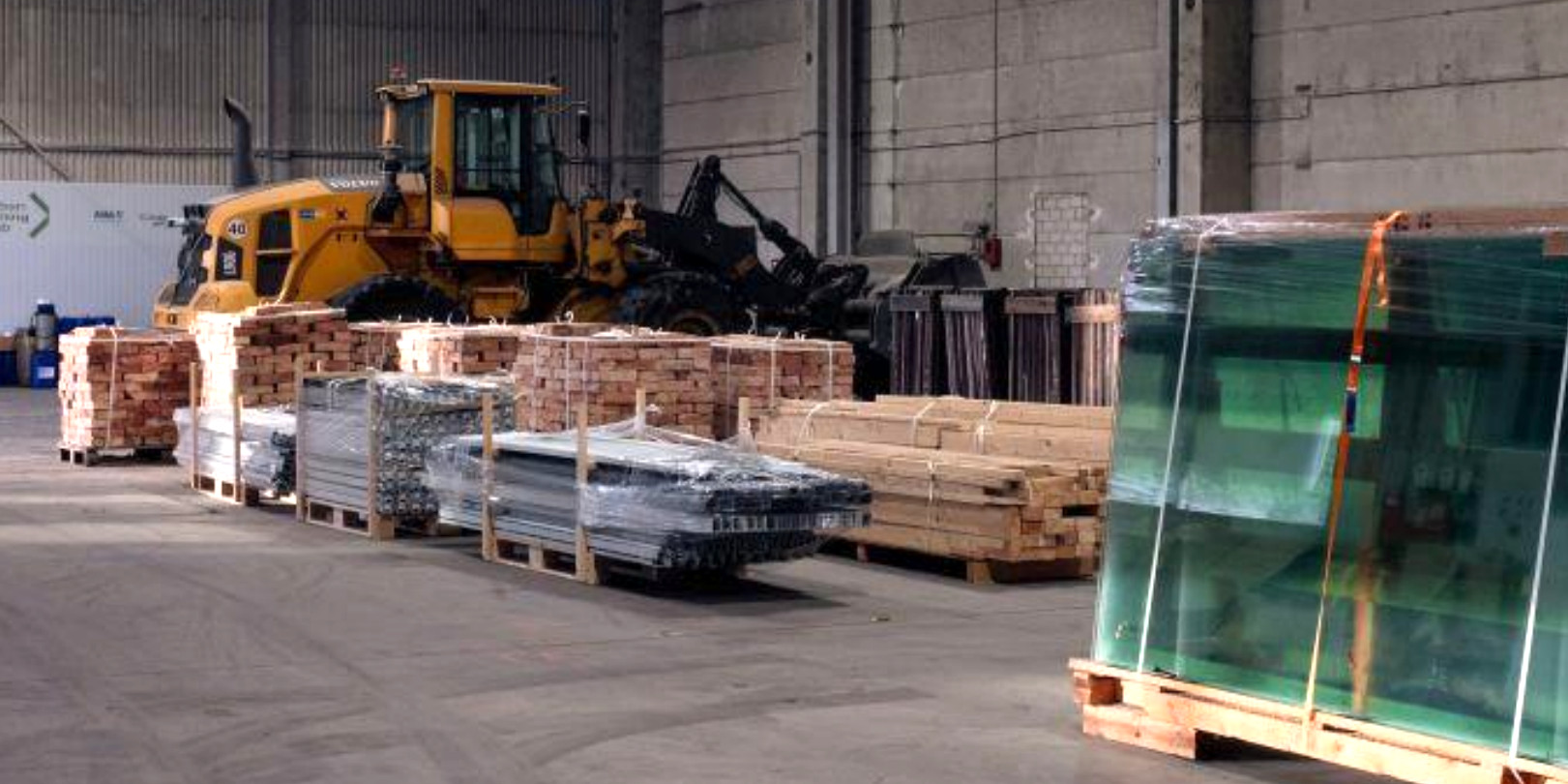 Innenansicht einer fensterlosen Industriehalle. Links steht eine Palette mit verpackten Glasscheiben, daneben Paletten mit Kanthölzern und Metallprofilen, dahinter Paletten mit Ziegeln. Im Hintergrund steht ein großer Radlader.