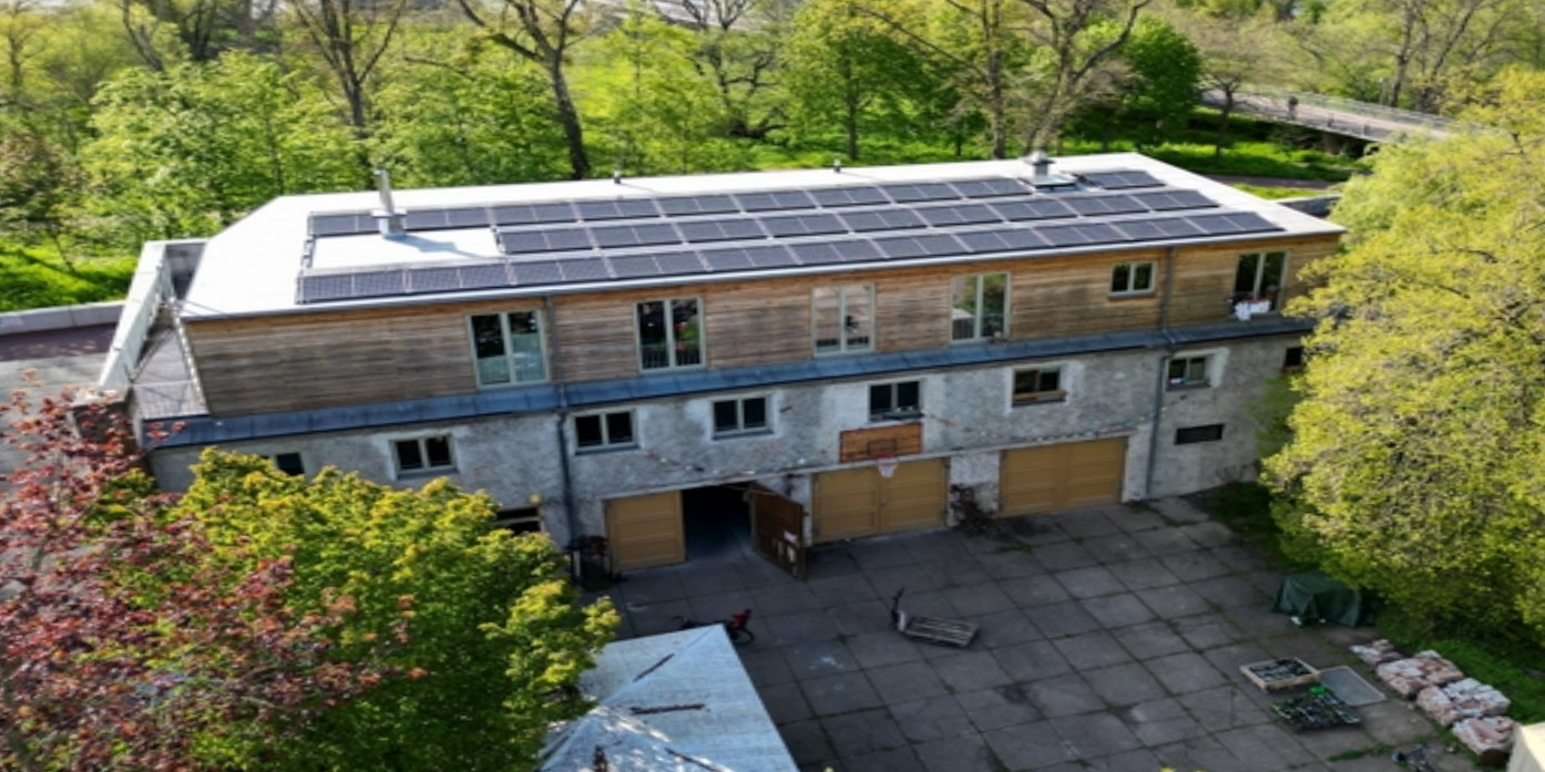 Luftbild eines zweigeschossigen Gebäudes mit weißem Putz, das mit einem Geschoss mit Holzfassade aufgestockt wurde. Auf dem Flachdach ist eine PV-Anlage installiert. Vor dem Gebäude ist ein gepflasterter Hof zu sehen, ringsum zahlreiche Bäume.
