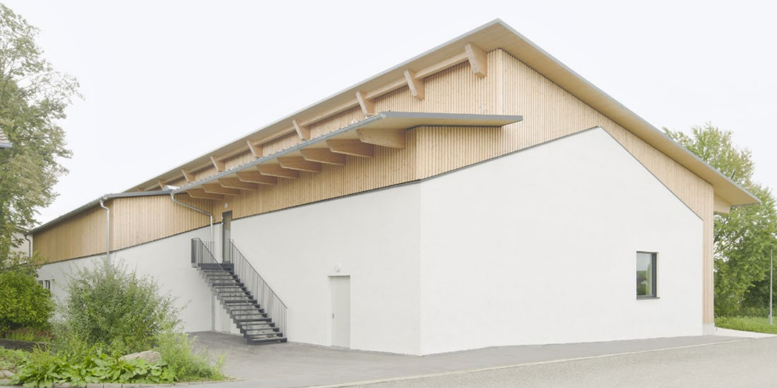 Eine Sporthalle mit versetztem Pultdach. In der Giebelfassade ist die Kubatur eines niedrigeren Gebäudes mit Satteldach durch eine weiße Putzfläche ablesbar. Die darüber gesetzte Dachkonstruktion setzt sich davon in hellem Holz ab.
